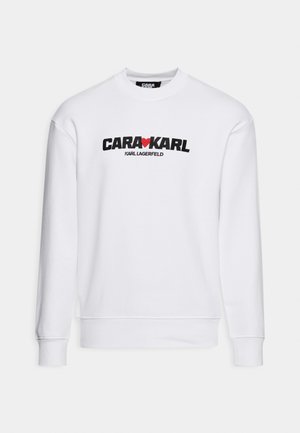 Biała bluza crewneck z długimi rękawami i czarnym napisem "CARA KARL" oddzielonym czerwonym sercem oraz mniejszym napisem "KARL LAGERFELD" poniżej.