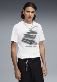 Maglietta bianca in cotone con una grafica di un'auto Porsche e logo, con maniche corte e scollo tondo, indossata con pantaloni neri.