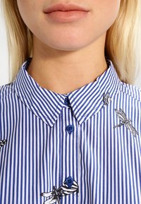 Blå och vit randig skjorta med knappar och spetsig krage, med svart libellbroderi och blå knappar fram.