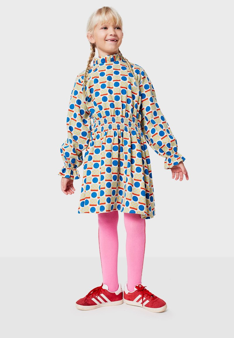 Abito colorato con vita arricciata, maniche lunghe e colletto alto, con cerchi blu e arancioni su uno sfondo chiaro. Collant rosa e scarpe rosse.