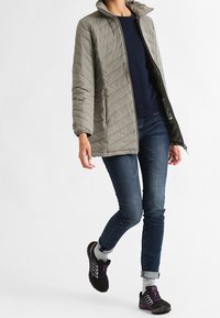 Light gray quilted jacket with a high collar, worn over a navy sweater. Paired with dark denim jeans and black athletic shoes with purple accents.