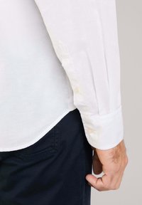 Witte lange mouwen shirt van lichtgewicht stof, met een enkelknop manchet en een gebogen zoom, gecombineerd met donkere broek.