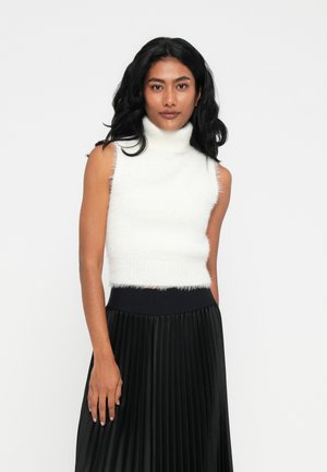 Donna con lunghi capelli neri che indossa un maglione bianco senza maniche con colletto alto e una gonna nera plissettata a vita alta, in piedi contro uno sfondo bianco.