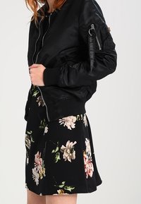 Svart bomberjacka med en blank yta, med ribbade ärmslut och en dragkedjeficka. Matchas med en svart kjol med blommönster.