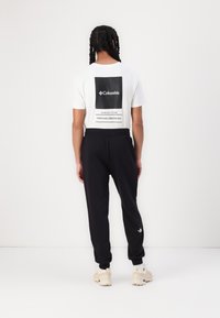 Hvid kortærmet t-shirt med en sort firkantet grafik, der har teksten "Columbia" og "Since 1938", parret med sorte sweatpants og beige sneakers.
