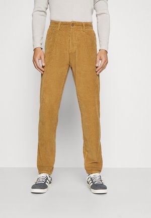 Mann trägt senfgelbe Cordhosen, weißes Langarmshirt und schwarz-weiße Sneakers, steht vor einem einfarbigen Hintergrund.