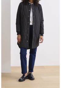 Schwarze, gesteppte lange Jacke mit Druckknopfverschluss, gerippten Bündchen und rundem Halsausschnitt, kombiniert mit dunkelblauen schlanken Hosen und schwarzen Loafern.