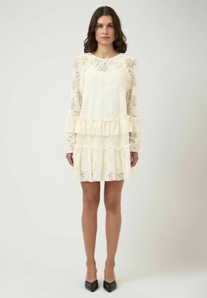 Femme debout, portant une robe longue en dentelle crème à manches longues avec des volants et des talons noirs pointus, devant un fond blanc uni.