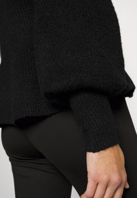 Gros plan d'une personne portant un pull noir texturé à manches longues bouffantes et un pantalon noir ajusté, montrant les détails du bras et de la hanche.