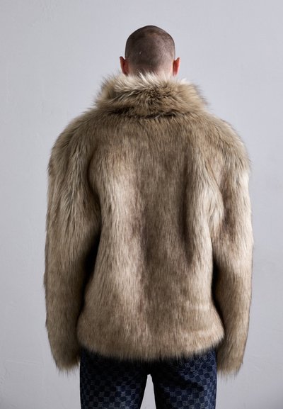 Faux fur jakke i lysebrun med en tæt, blød tekstur. Har en høj krave og en let løs pasform, parret med mønstrede mørke jeans.