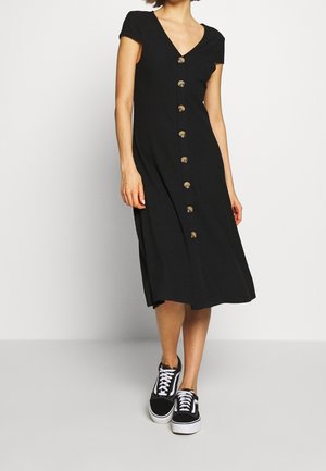 Czarna prążkowana sukienka midi z krótkimi rękawami, dekoltem w serek i mosiężnymi guzikami z przodu. Połączona z czarnymi trampkami.