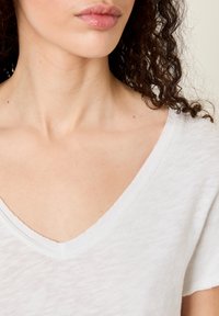 Närbild av en persons hals och nedre ansikte med lockigt hår, iförd en vit V-ringad topp mot en neutral bakgrund.