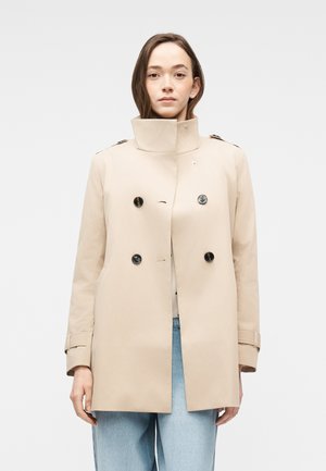 Kvinde iført en beige dobbeltradet trenchcoat med sorte knapper og lyseblå jeans, stående mod en ensfarvet lys baggrund.
