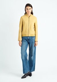 Veste bomber en cuir jaune avec fermeture éclair à l'avant, col plat et manches longues, associée à un jean large bleu clair et des bottes noires.