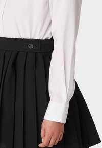 Falda negra con plisados, cinturilla de tela y detalles de botones, combinada con una camisa blanca de manga larga con puños abotonados.
