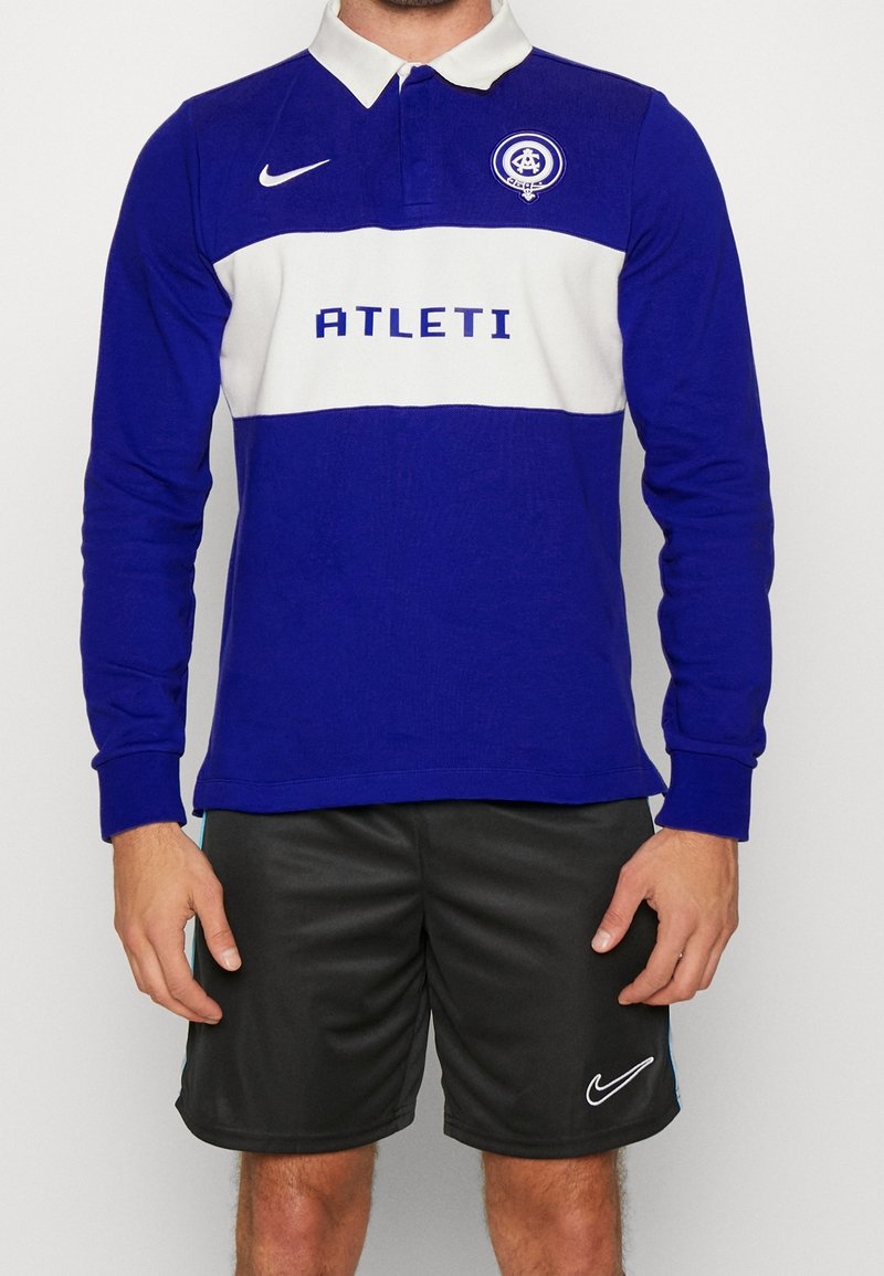 Blå rugbytröja med lång ärm och vit horisontell rand samt texten "ATLETI". Har krage och Nike-logotyp. Kombineras med svarta shorts.