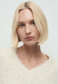 Suéter de manga corta, color crema, suave y peludo, con escote en V. La modelo tiene el cabello lacio, hasta los hombros, de color rubio y un maquillaje natural.