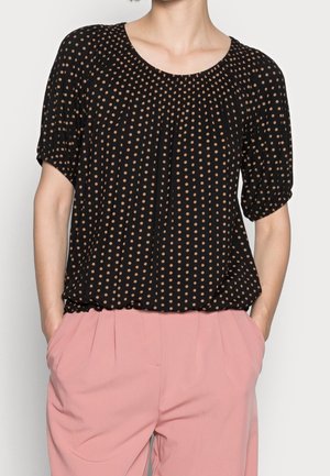 Femme portant un chemisier noir à manches courtes avec des pois beiges et un pantalon plissé rose taille haute, les mains dans les poches.