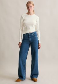 Młoda kobieta stoi, ubrana w białą bluzkę z długim rękawem, niebieskie jeansy z wysokim stanem i szerokie nogawki, oraz brązowe zamszowe buty na tle jednolitym.