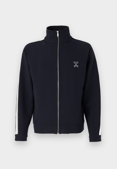Sweatshirt zippé bleu marine avec un col montant, doté de rayures latérales blanches et d'un logo brodé sur la poitrine. Tissu doux et poignets côtelés.
