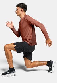 T-shirt de sport rouge à manches longues associé à un short noir. Le coureur porte des chaussures noires et blanches, montrant un mouvement dynamique vers l'avant.