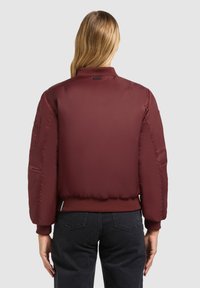 Bordeaux bomberjack met een gladde textuur, ribbelmanchetten en -zoom, en zijzakken; heeft een subtiel logo bij de kraag.