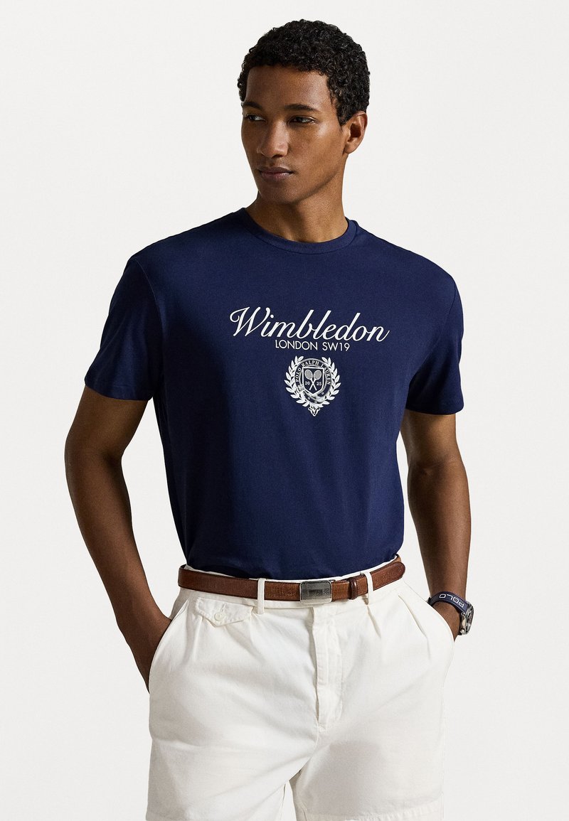 Tricou din bumbac bleu marin cu text alb "Wimbledon", logo cu scut, mâneci scurte, guler rotund, asociat cu pantaloni scurți albi și o centură maro.