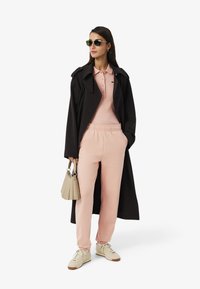 Trench nero, polo rosa e pantaloni tuta, scarpe da ginnastica beige, occhiali da sole verdi e una borsa beige chiaro con dettagli plissettati.