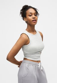 Νεαρή γυναίκα με σγουρά μαλλιά, φορώντας γκρι αμάνικο crop top και ανοιχτό γκρι φόρμα, στέκεται μπροστά σε λευκό απλό φόντο.