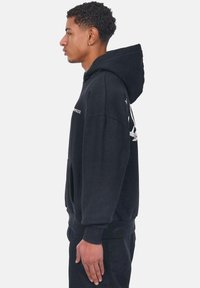 Schwarzer oversized Hoodie mit einer vorderen Kängurutasche, der aus einem strukturierten Stoff gefertigt ist und eine Kapuze hat. Logo auf der Vorderseite und dem Ärmel bedruckt.