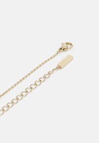 Gouden ketting met een kreeftsluiting en een rechthoekig label gegraveerd met "LACOSTE." Bevat afwisselende schakels en een gepolijste afwerking.