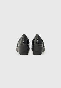 Chaussures en cuir verni noir avec un talon compensé, présentant une texture lisse et un logo de marque discret sur le talon, conçues pour le confort.
