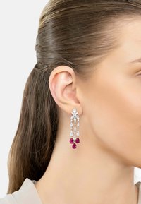 Woman's ear with dangling silver earrings featuring clear and red teardrop-shaped gemstones.