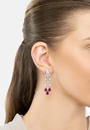 Woman's ear with dangling silver earrings featuring clear and red teardrop-shaped gemstones.