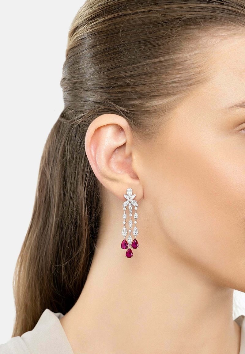 Woman's ear with dangling silver earrings featuring clear and red teardrop-shaped gemstones.