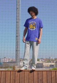 Tricou din bumbac albastru cu logo-ul echipei Golden State Warriors, asociat cu pantaloni din denim deschis și teniși albi, stând lângă un gard din plasă.