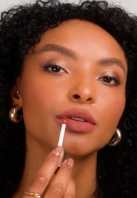 Nainen kiharat mustat hiukset ja kultaiset renkkikorvakorut levittämässä vaaleanpunaista huulikiiltoa huulilleen sisätiloissa neutraalin taustan edessä.