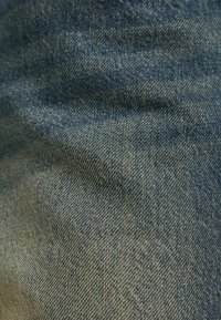 Denim-kangas, jossa vaihtelevat siniset sävyt ja sileä pinta, esittäen vinokudonta kuvioita ja hienovaraisia valohaalistumia joillakin alueilla.