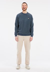 Felpa crewneck in pile di colore blu scuro con polsini e orlo a costine, abbinata a pantaloni beige a gamba dritta e sneakers chiare.