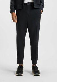Black athletic joggers with a tapered fit, made from soft fabric. Features two side pockets and elastic cuffs. Worn with black sneakers.