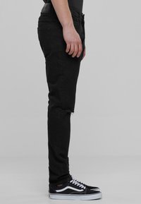 Jeans ajustados negros con dobladillo desgastado, hechos de denim, con un diseño clásico de cinco bolsillos. Llevados con zapatillas en blanco y negro.