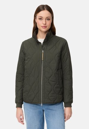 Kobieta w ciemnozielonej pikowanej kurtce na zamek i jasnoniebieskich dżinsach, stojąca na tle jednolitego, jasnego tła.