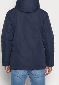 Marinblå jacka med huva, med en slät, matt yta och rak passform. Har en fåll vid midjan och långa, något avsmalnande ärmar.