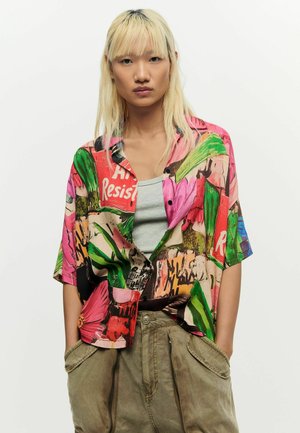 Tânăr cu păr lung și blond purtând o cămașă colorată cu imprimeu abstract peste un maieu gri și pantaloni khaki largi, cu mâinile în buzunare.