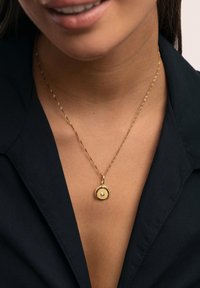Collar de oro con cadena retorcida y colgante circular que presenta una piedra preciosa central. Llevado sobre el escote de una camiseta negra.
