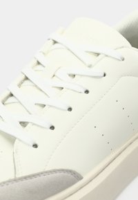 Sneakers en cuir blanc avec des lacets blancs, un embout en daim gris, de petites aérations sur les côtés et une semelle de couleur crème.