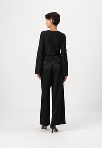 Top nero con texture, maniche lunghe, abbinato a pantaloni a gamba larga in satin nero a vita alta. Modella che indossa tacchi neri, di spalle rispetto alla fotocamera.