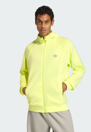Junger Mann mit lockigem Haar, der einen neongelben Zip-Hoodie mit Adidas-Logo und beige Hose trägt, Hände in den Taschen, neutraler Hintergrund.