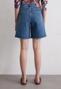 Jeansshorts in einem mittleren Blauton mit geradem Schnitt, gesäumten Kanten und zwei Gesäßtaschen. Tragen mit schwarzen Sandalen.