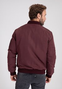 Giubbotto bomber bordeaux con colletto e polsini a coste, dotato di una superficie liscia e orlo elastico. Dettagli visibili sulle cuciture laterali.